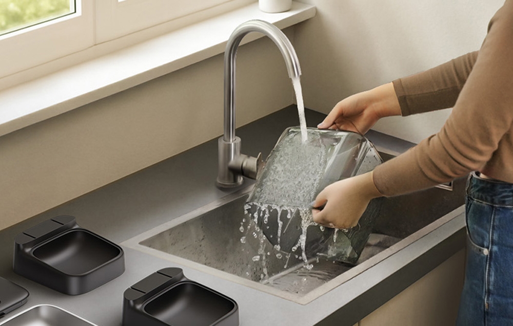 A woman washes the components of a food dispenser under running water.