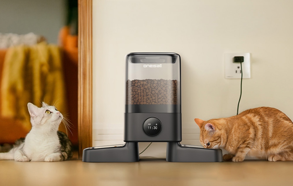 A ginger cat uses the food dispenser. A white and gray cat lies nearby.