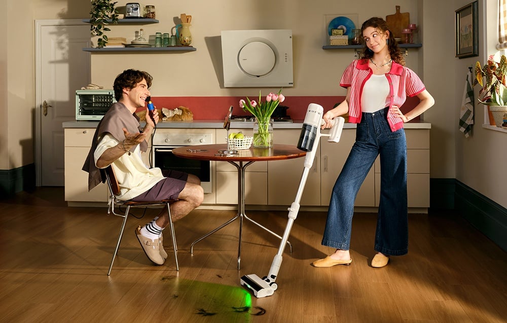 Mężczyzna siedzi przy stole w kuchni i śmieje się, a kobieta obok odkurza podłogę bezprzewodowym odkurzaczem.