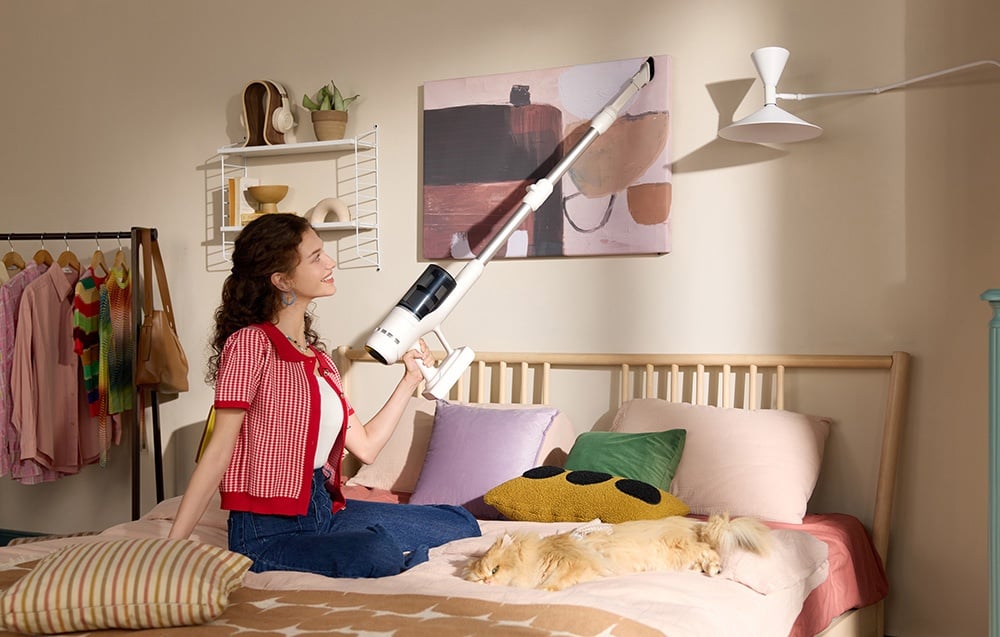 Kobieta siedzi na łóżku i odkurza górną część ściany za pomocą odkurzacza bezprzewodowego, a na łóżku leży śpiący kot.