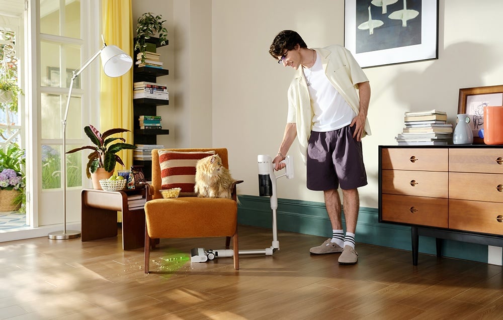 Mężczyzna odkurza podłogę bezprzewodowym odkurzaczem pod fotelem, na którym siedzi kot.