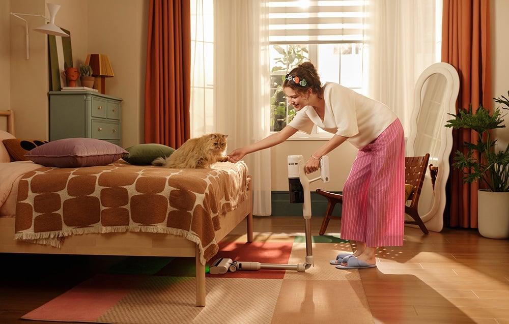 Kobieta w różowych spodniach w paski odkurza podłogę przy łóżku i głaszcze kota siedzącego na łóżku.