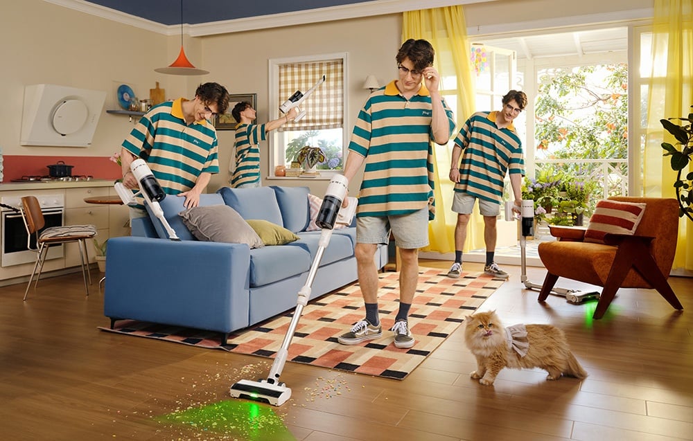 Mężczyzna w zielono-beżowej koszulce widoczny w kilku ujęciach jednocześnie sprząta salon odkurzaczem bezprzewodowym, podczas gdy kot w sweterku siedzi na podłodze.