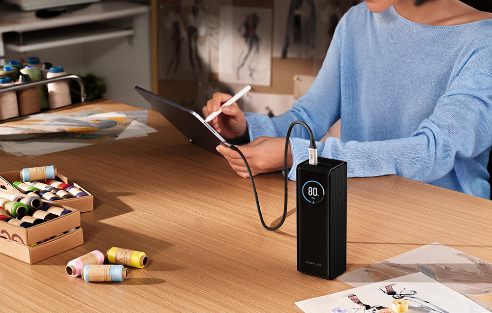 A woman in a blue sweater draws on a tablet at a wooden table with a power bank and spools of colorful threads