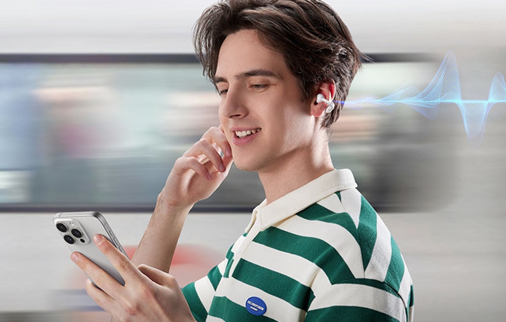 A man in a green and white T-shirt uses a phone and touches the receiver in his ear, standing against the background of a passing train