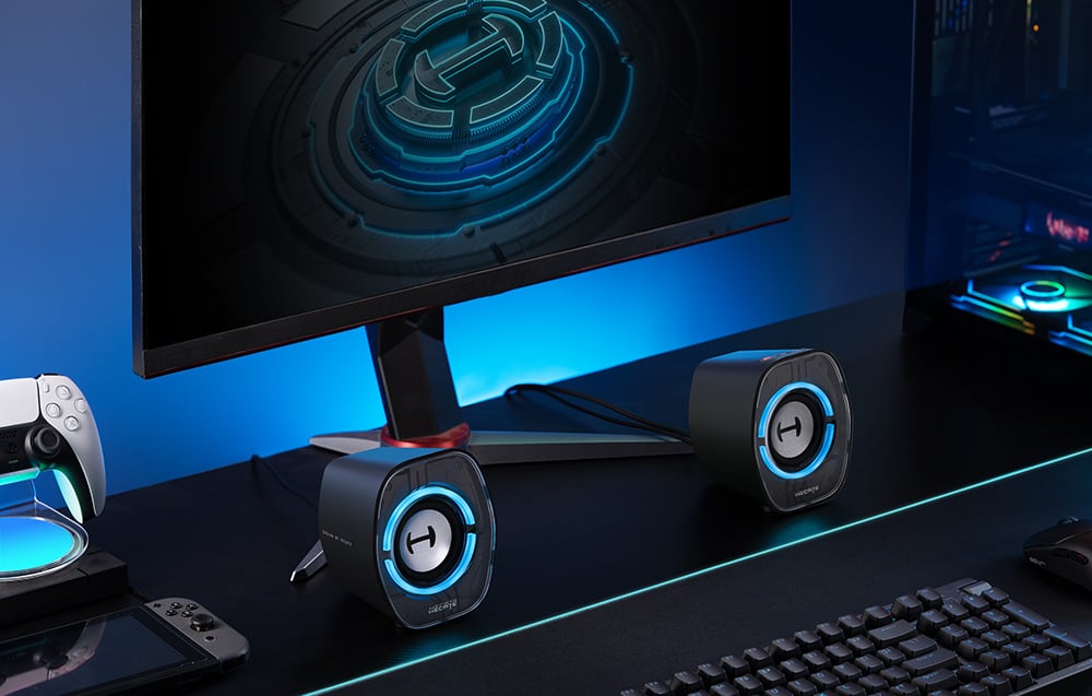 Černé reproduktory Hecate leží na stole vedle monitoru, klávesnice a herní konzole.