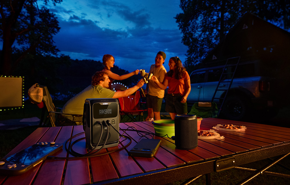 A group of people toast at a camping table with a power station and speaker in the evening