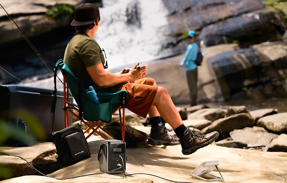 A person sitting on a camping chair by a waterfall with a portable power station on the ground