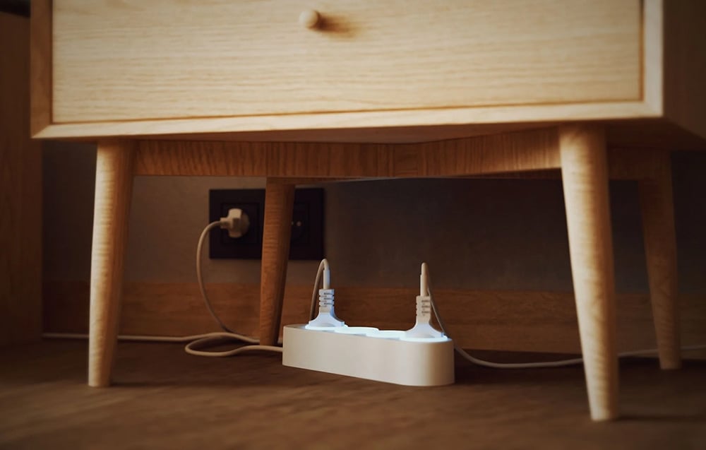 A white Shelly power strip under a wooden bedside table with two devices plugged into it