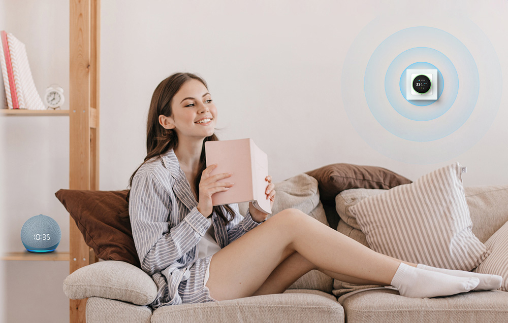 A young woman sits on a couch and holds a notebook. There is a thermostat on the wall and a voice assistant on the shelf