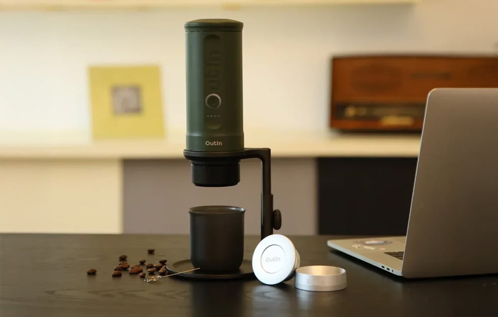 Elektryczny zaparzacz do kawy Outin w czarnym kolorze ustawiony na podstawce nad kubkiem na biurku obok laptopa i rozrzuconych ziaren kawy