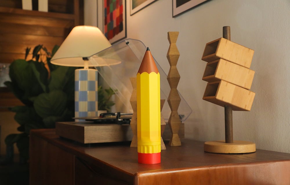 Duży przedmiot w kształcie żółtego ołówka stoi na komodzie obok designerskiej lampy i drewnianych dekoracji