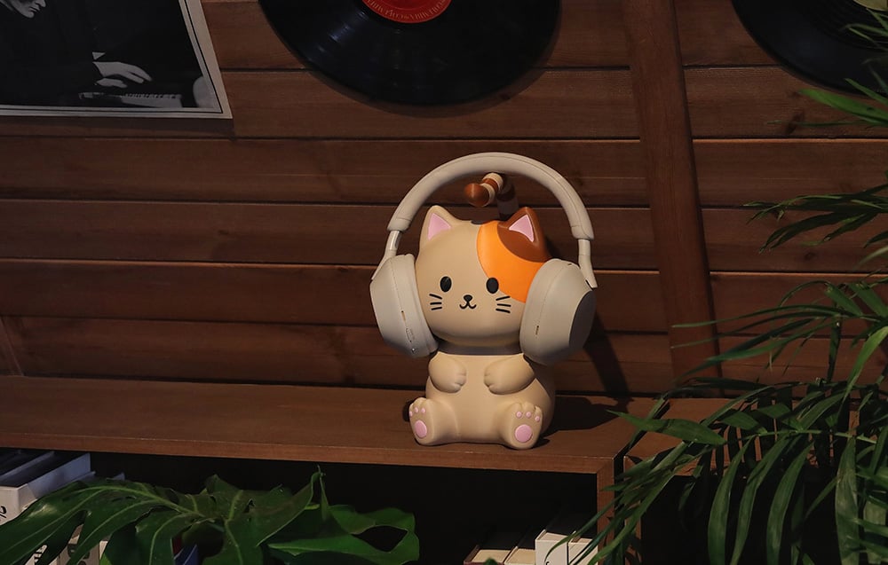 Figurka kota z pomarańczową łatką ma na uszach duże słuchawki i stoi na półce przy roślinach
