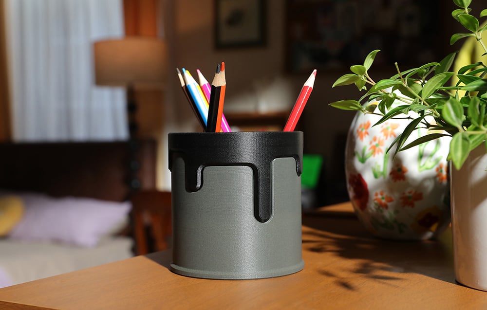 Grafitowy pojemnik na ołówki z ozdobnym wzorem trzyma kolorowe kredki i stoi obok doniczki z rośliną