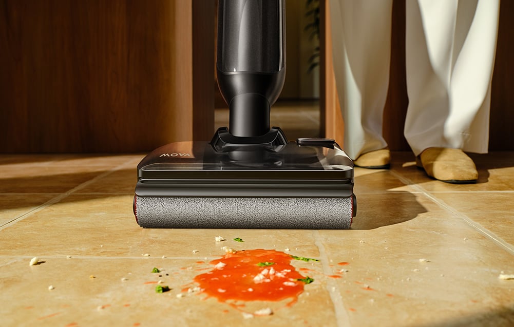 Čierna hlava umývacieho vysávača s rotujúcim valčekom stojí na podlahe vedľa červenej škvrny od omáčky