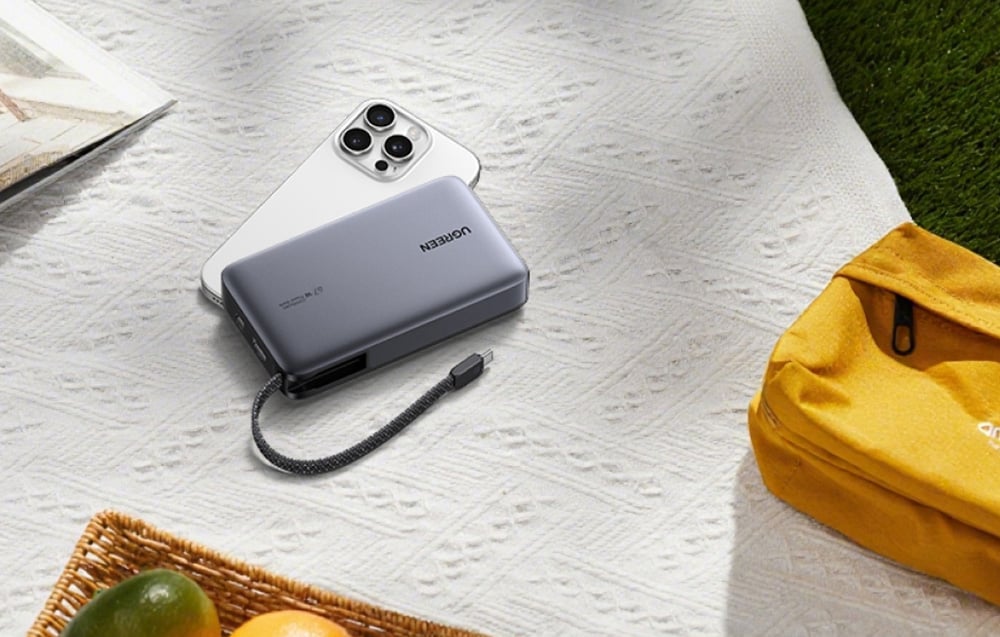 Egy Ugreen power bank és egy iPhone egy piknik takarón fekszik egy sárga táska mellett, utalva a termék utazás és szabadtéri használatára.