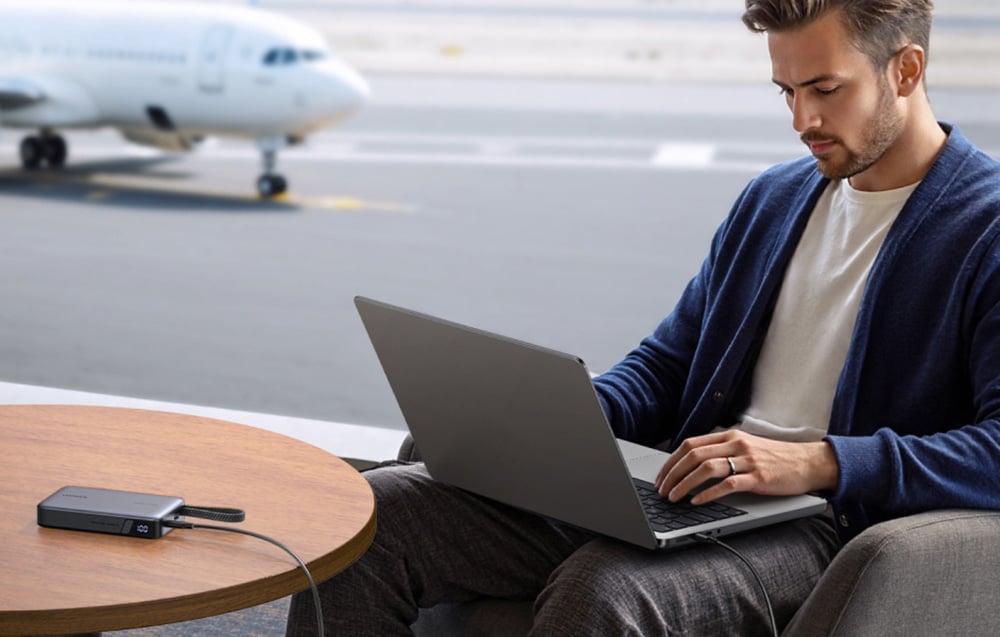 Egy férfi laptopon dolgozik egy repülőtéri terminálon, és az eszközt egy Ugreen power bankról tölti. A háttérben egy repülőgép látható.