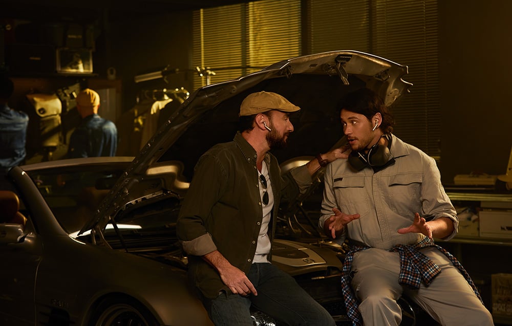 Dois mec�nicos est�o sentados em um carro com o cap� aberto, ambos usando fones de ouvido com fun��o de tradu��o.