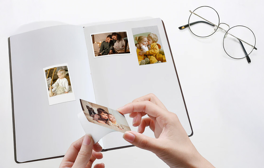 A person pasting a photo of children into a photo album, glasses standing next to it