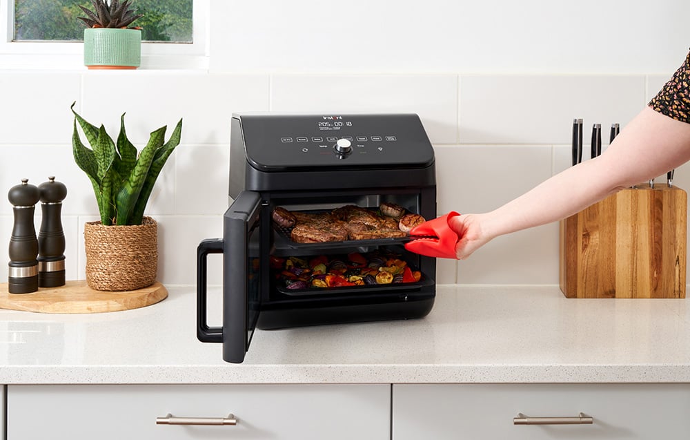 Ręka w czerwonej rękawicy wysuwa blaszkę z grillowanym mięsem i warzywami z piekarnika