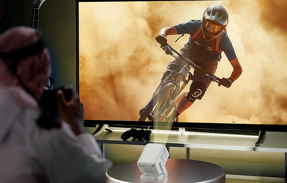 A projector placed on a table displays a dynamic image of a mountain biker riding in clouds of dust on a TV screen
