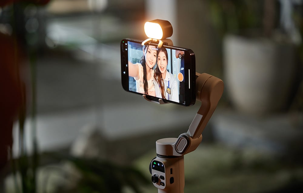 A smartphone mounted in a beige gimbal with a flash on, displaying a selfie of two women.