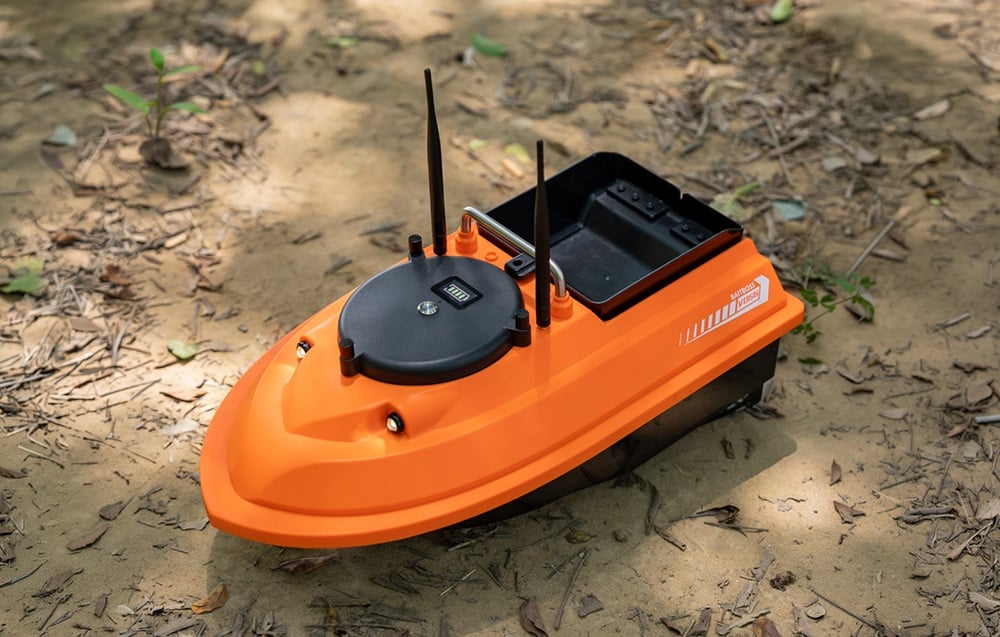 Orange bait boat with two antennas and a handle, standing on a sandy shore.