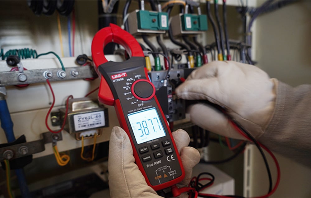 UNI-T digital clamp meter held in an electrician's glove during testing in a control cabinet.