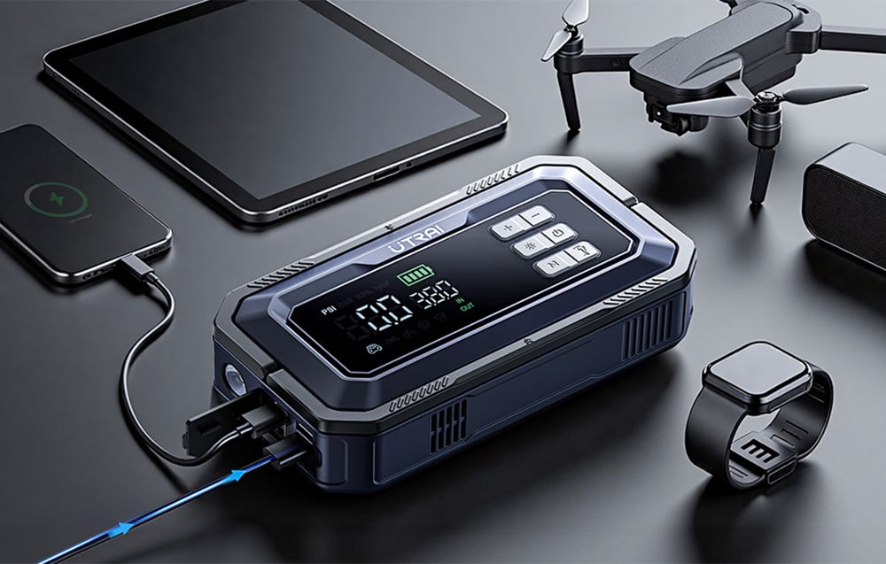 A car jump starter on a table charges a smartphone and tablet next to a watch and a drone.