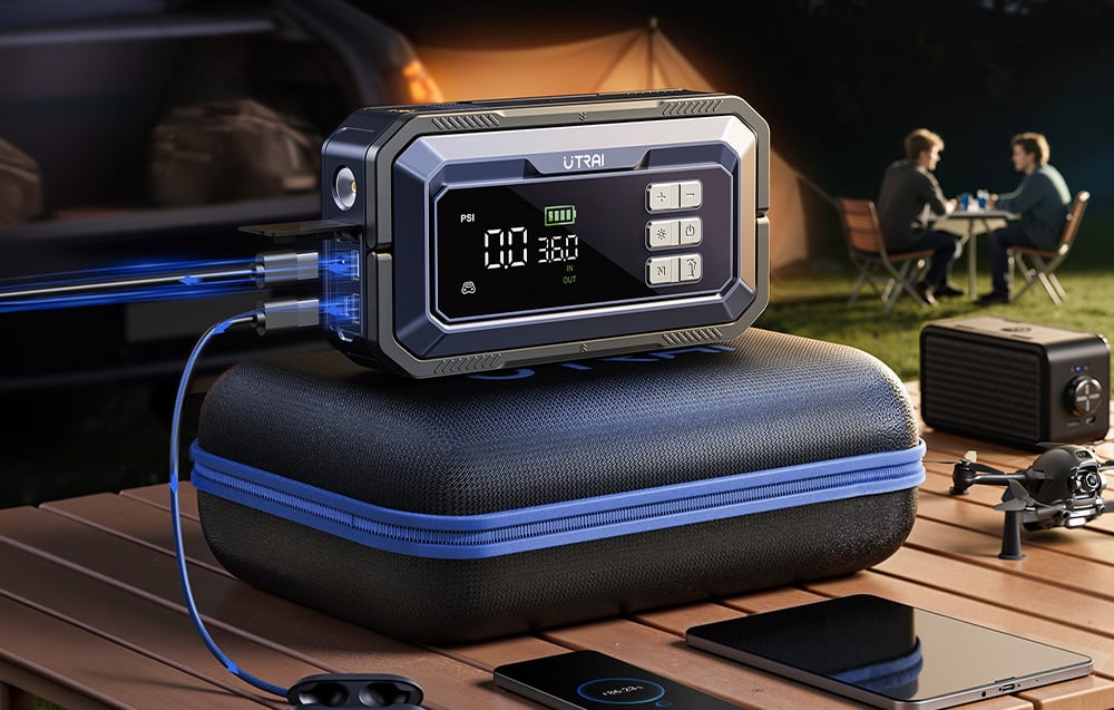 A car jump starter on a suitcase is charging a phone against a backdrop of a tent and people talking.