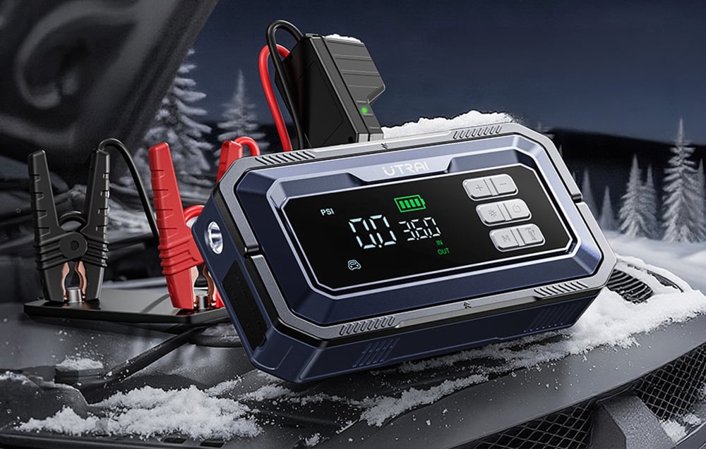 A car jump starter with jumper cables attached lies on a snow-covered car hood.
