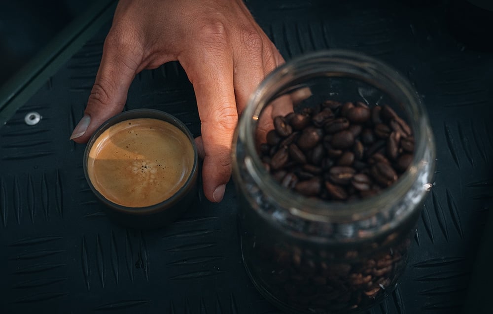 Close-up of a hand with a small cup of coffee with foam, next to it is a jar filled with coffee beans