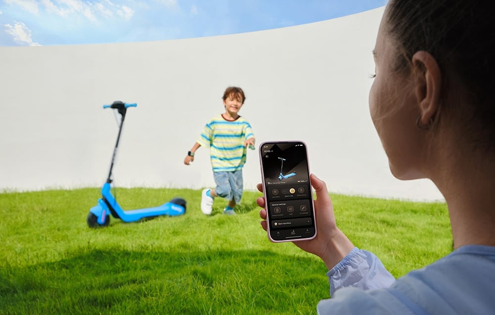 Μια γυναίκα χρησιμοποιεί την εφαρμογή στο smartphone της, ενώ ένα παιδί τρέχει δίπλα σε ένα παρκαρισμένο σκούτερ στο γρασίδι