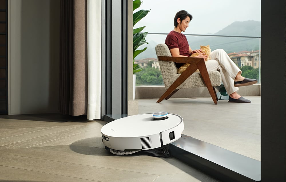 Robot sprzątający przejeżdża przez niski próg balkonowy, podczas gdy mężczyzna siedzi na tarasie z kotem na kolanach