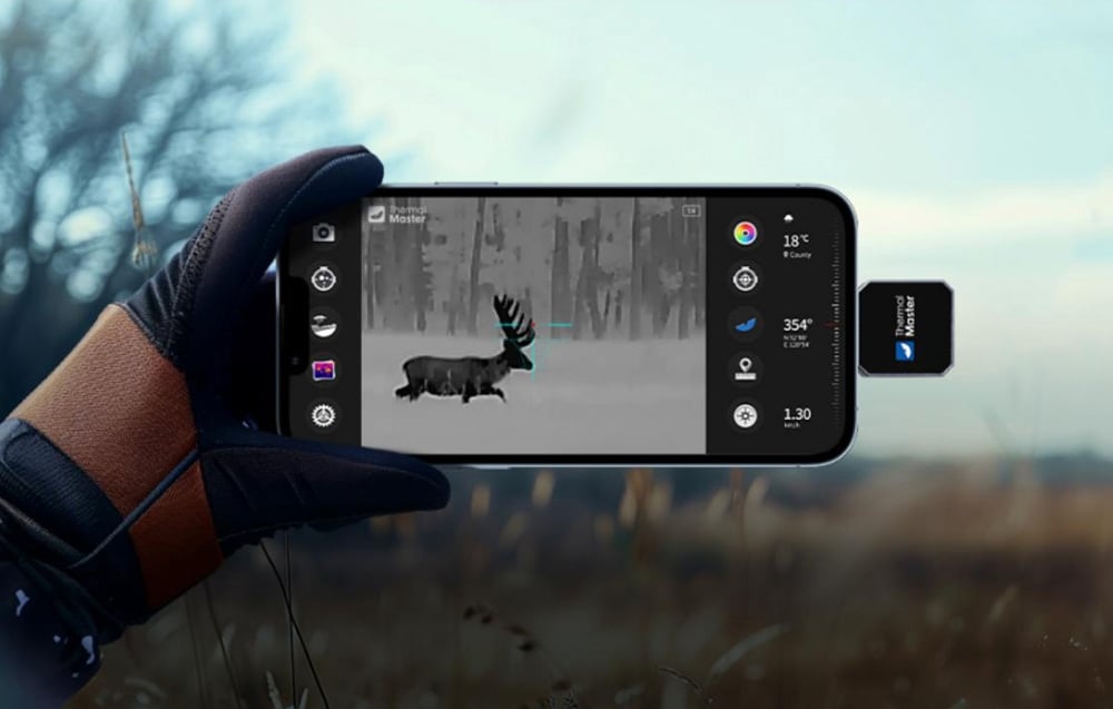 A smartphone connected to a small thermal imaging camera displays a black-and-white image of a deer in a clearing.