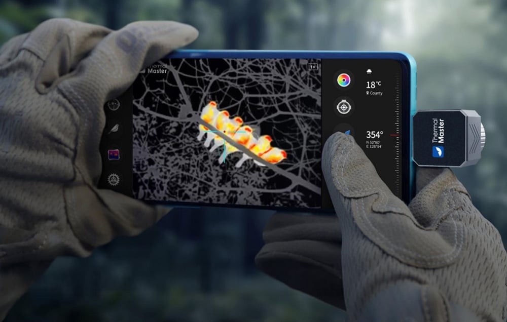 The screen of a phone with a thermal imaging camera displays a color thermal image of a caterpillar hidden in the branches.