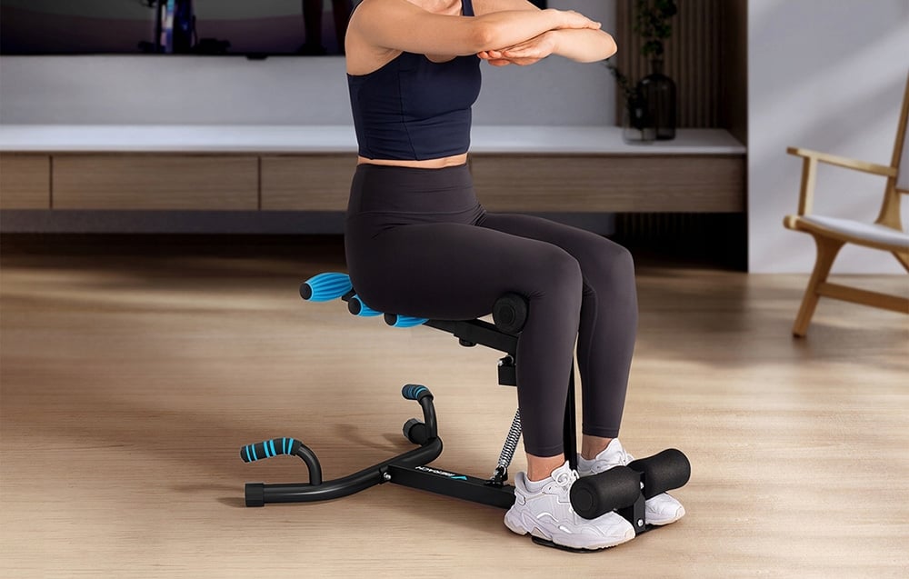 A woman with her arms crossed sits on a black machine in a living room