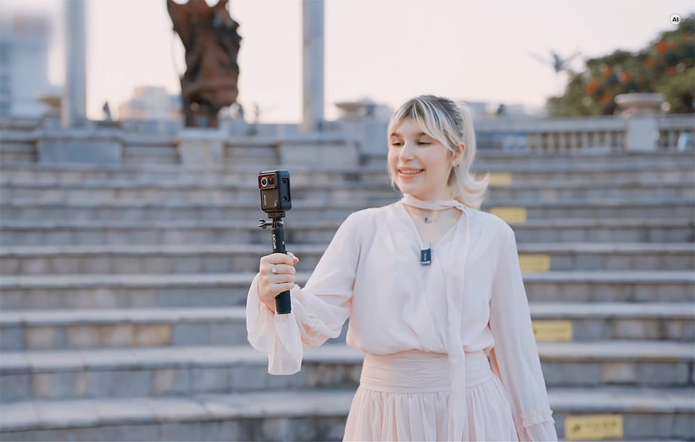 Szőke nő, aki városi környezetben egy szelfibotra rögzített SJCAM akciókamerát tart a kezében.
