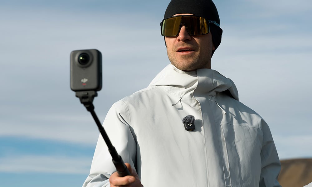 Mężczyzna w białej kurtce, czapce i goglach przeciwsłonecznych nagrywa vloga kamerą na wysięgniku